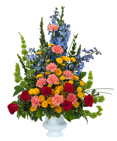 Bright sympathy arrangement in coral, blue, red and yellow with greenery in a white urn. 