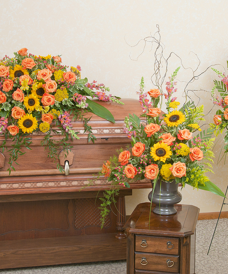 Three funeral flower arrangements: A casket spray, standing spray and urn design in sunset colors. 