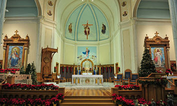 An ornate church altar displays religious statues, paintings, and Christmas decorations, surrounded by floral arrangements and pews in a spacious, vaulted room adorned with religious artwork and archi
