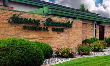 Hanson-Runsvold Funeral Home sign is mounted on a brick wall with neatly trimmed bushes and colorful flowers in front, accompanied by a building with a green roof and cloudy sky above.